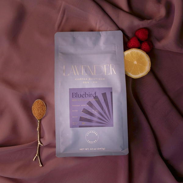 Lavender coffee package with a spoonful of coffee, lemon, and raspberries on a purple fabric background